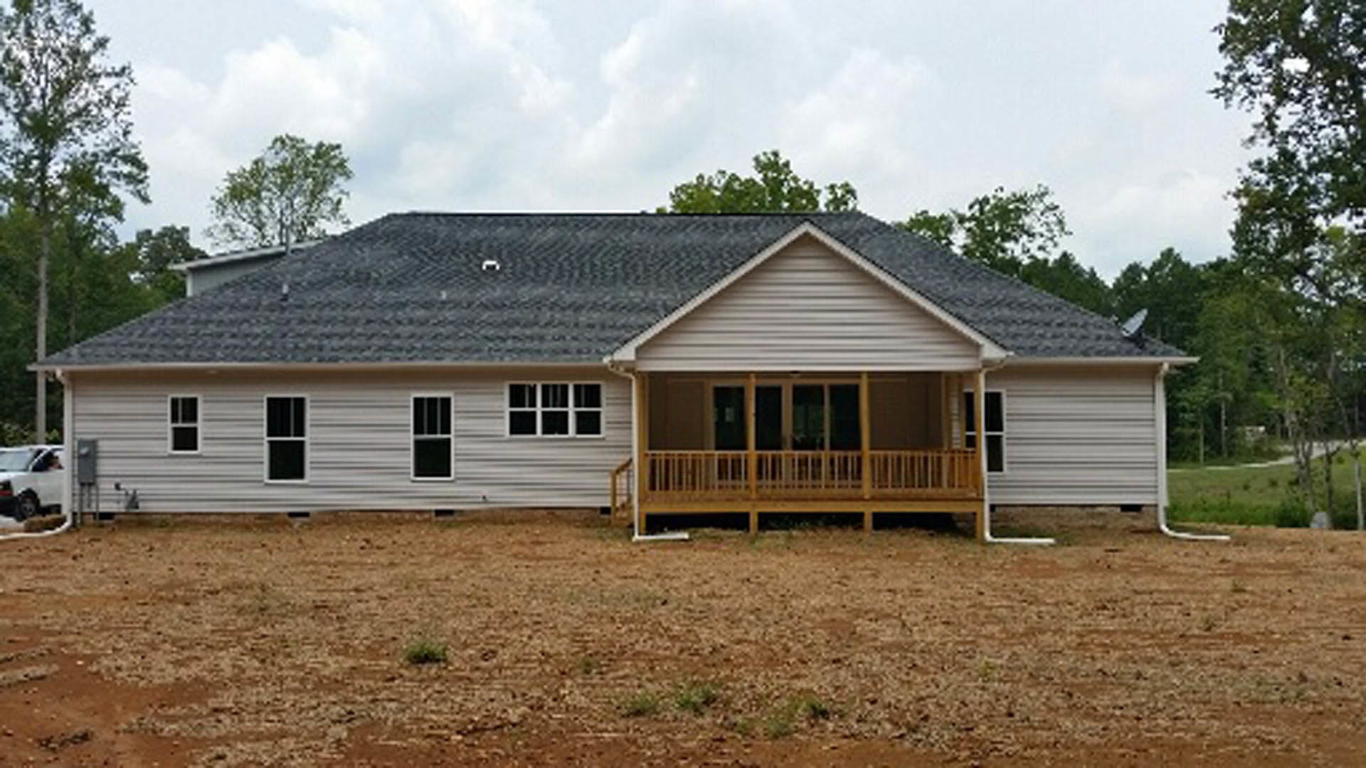 Front porch with white columns, light-colored siding, white-framed windows, dirt and grass yard, partially visible white car, cloudy sky overhead