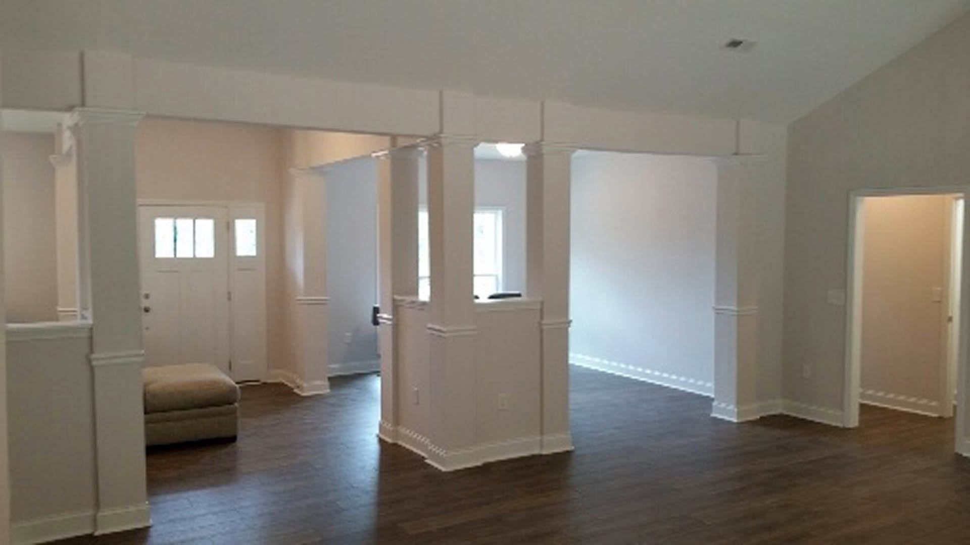 Living room with white plaster walls, light laminate flooring, white couch, upholstered footstool, and white door illuminated by overhead lighting