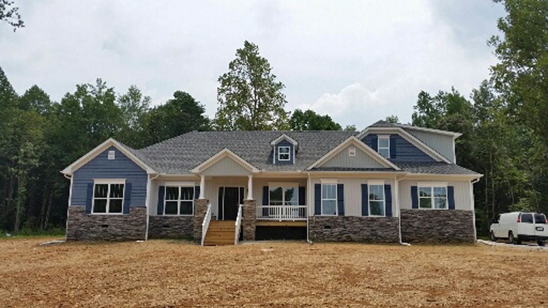 Two-story white house with covered porch, white railings, and stairs, set on a large grassy yard; white van parked nearby, dirt field with a person in the background, mature trees