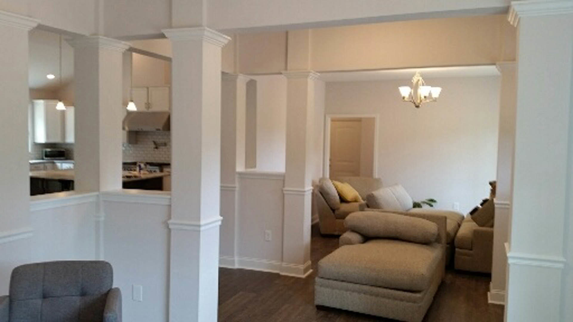 Open-concept living room with light-colored couch and accent pillows, hardwood flooring, and adjacent modern kitchen with white cabinetry