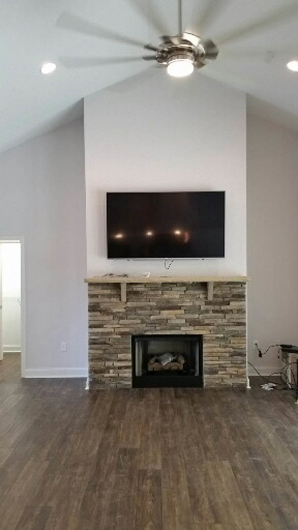 Stone fireplace with mounted TV above, wood flooring, neutral walls, and ceiling fan in a spacious living room.