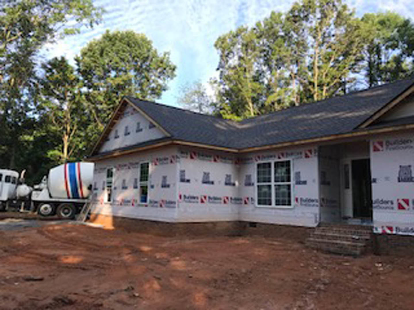 Two-story home with white stucco walls, large windows, and a cement mixer truck parked on dirt driveway; leafy tree and blue sky in background.