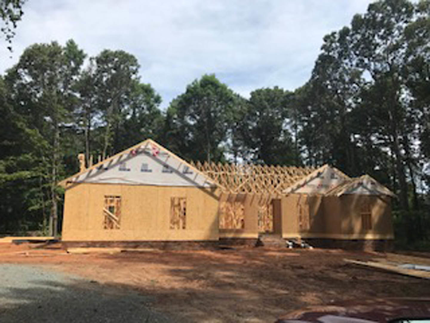 Framed house under construction with exposed wooden beams, partially finished roof, surrounded by tall trees and dirt ground