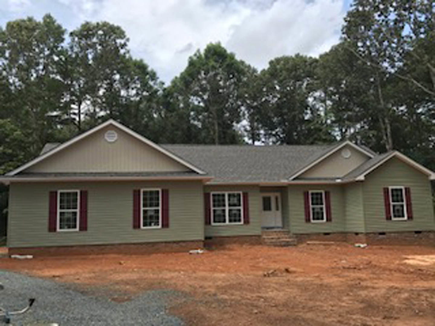 Partially built house with exposed framing, white door and windows, surrounded by trees and dirt construction area