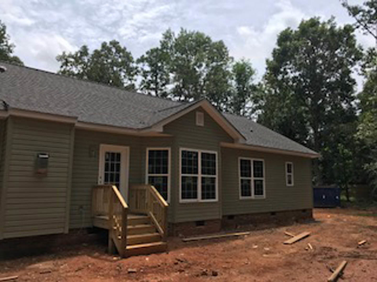 Partially built house with exposed wooden framing, outdoor wooden staircase, white-framed multi-pane windows, and trees in the background