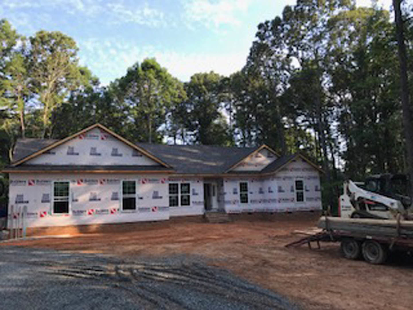 Two-story home under construction with exposed framing, construction truck parked on dirt driveway, mature trees surrounding site, cloudy sky overhead
