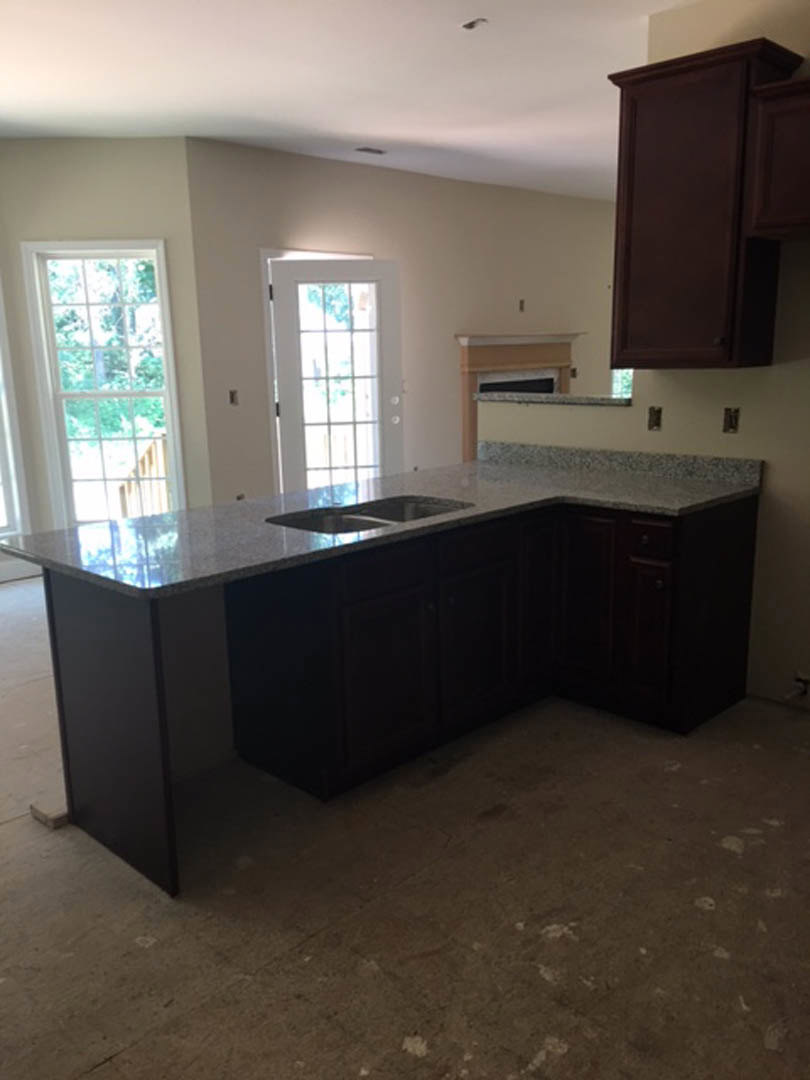 Granite kitchen countertop with stainless steel sink, brown cabinetry, window overlooking trees, hardwood flooring, and partial view of a fireplace