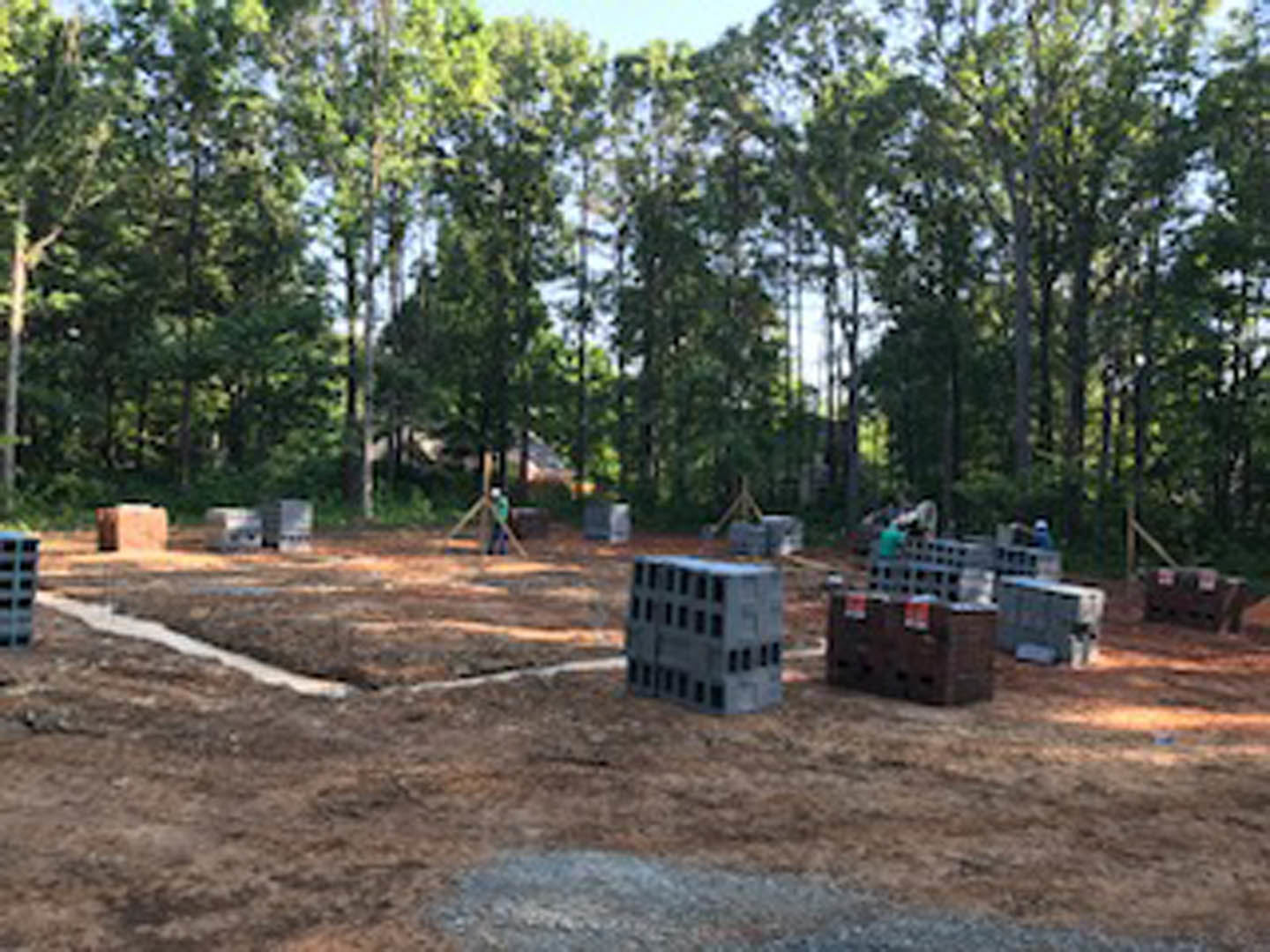 Cement blocks stacked on dirt ground at residential construction site, surrounded by trees and plants under open sky