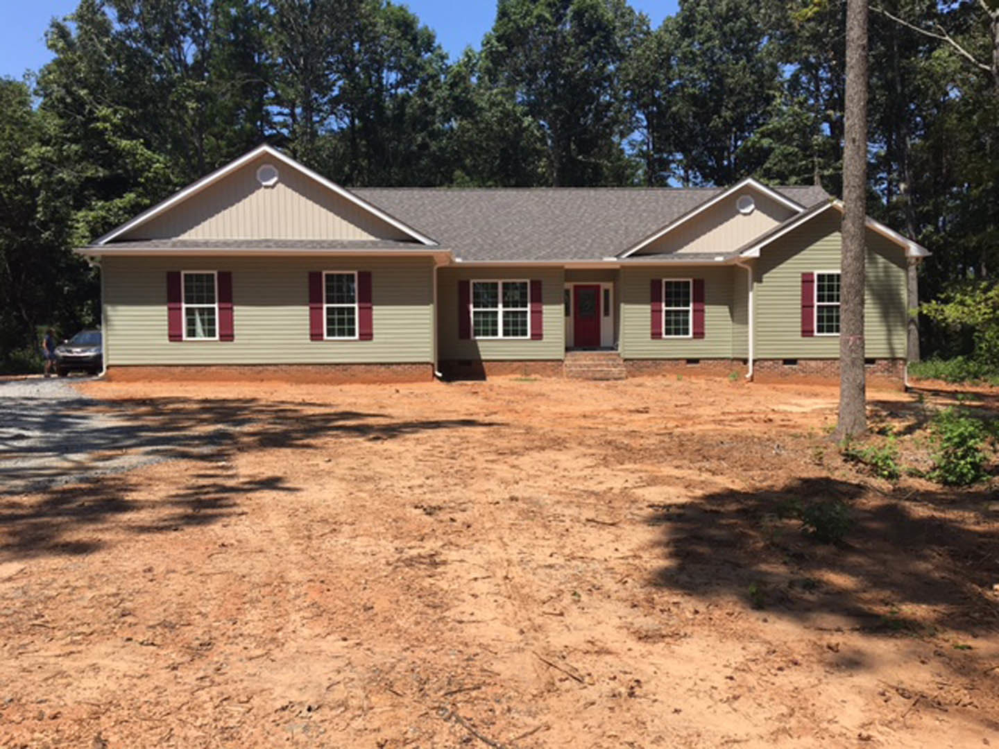 Two-story house with red shutters and red front door, white trim around windows, dirt yard in foreground, group of trees in background.