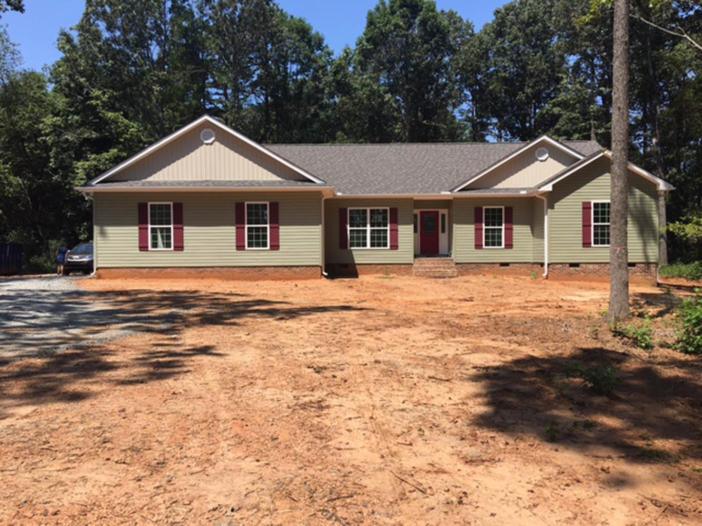 Two-story house with white siding, red front door, large windows, and covered porch, set on a dirt lot with scattered trees and a parked car in the background