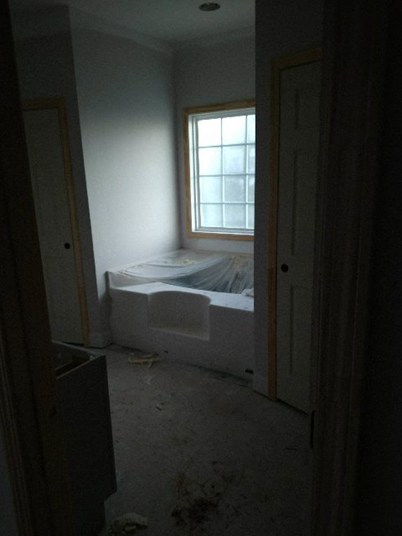 White freestanding bathtub with protective plastic covering in a plaster-walled room, large window, unfinished ceiling, and door visible.