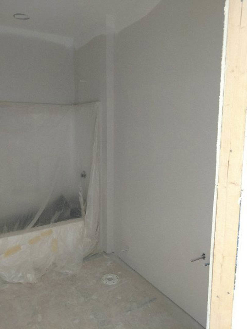 Freestanding bathtub wrapped in protective plastic, light plaster walls, concrete floor, recessed ceiling, unfinished bathroom remodel