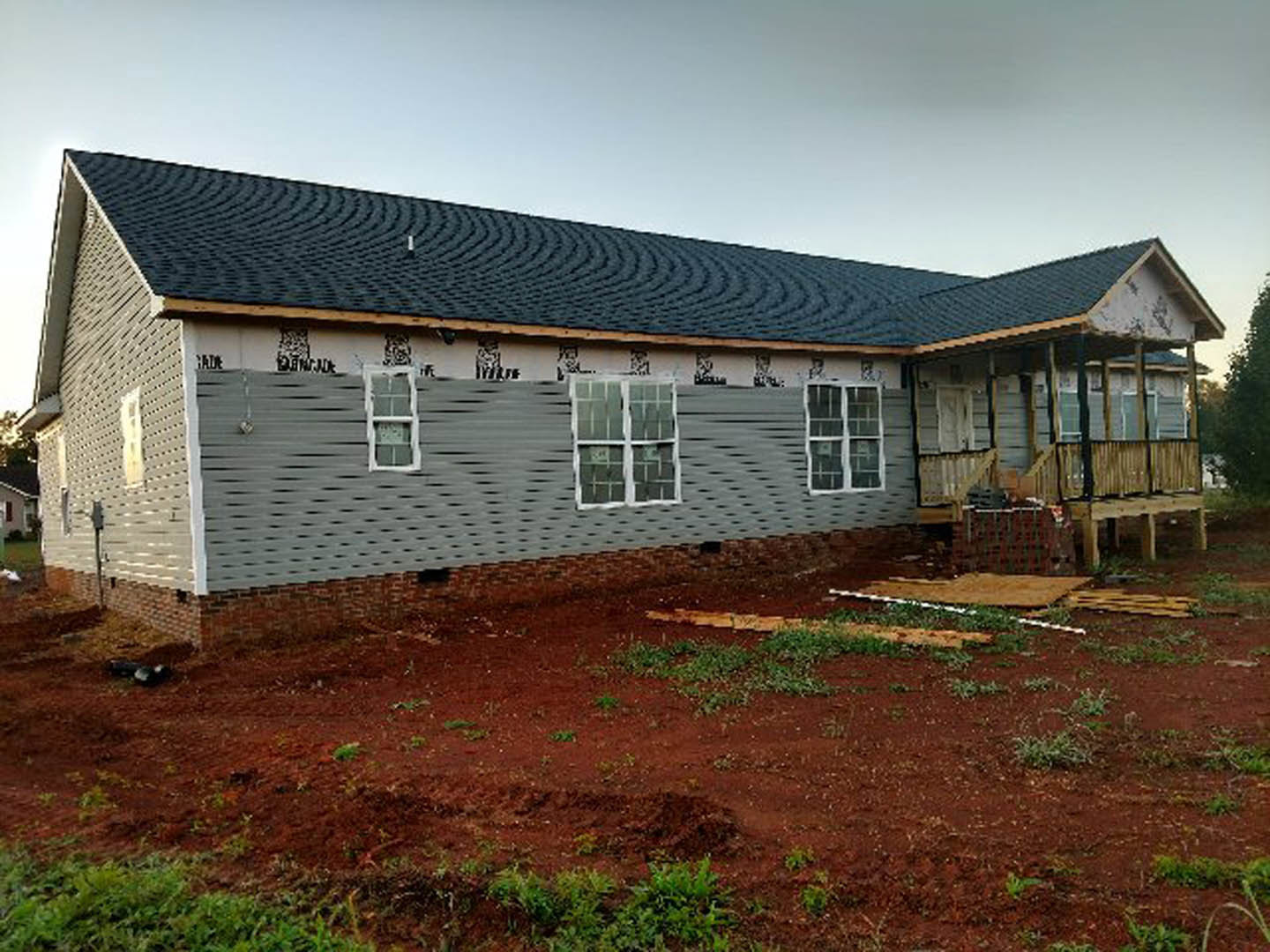 Single-story cottage with light siding, multiple windows, and a gray roof, set on a red dirt yard with sparse grass and plants under a clear sky