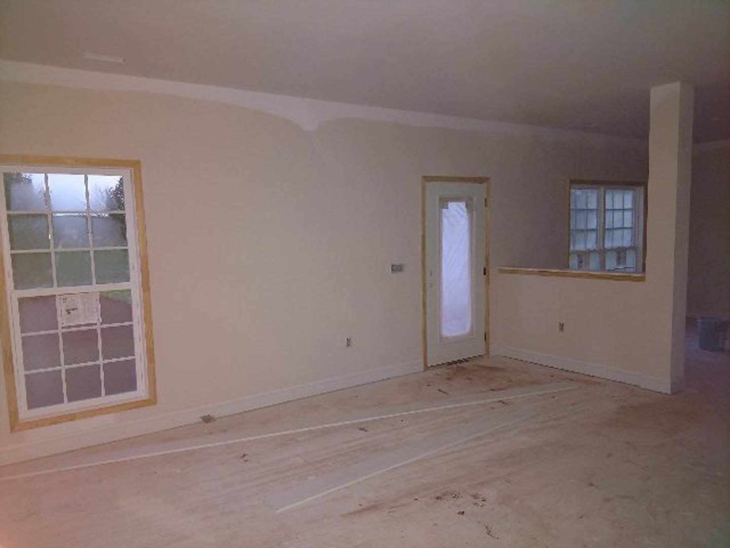 Spacious room with light laminate flooring, white plaster walls, large windows, and a modern door; white tape and paper visible on the floor.