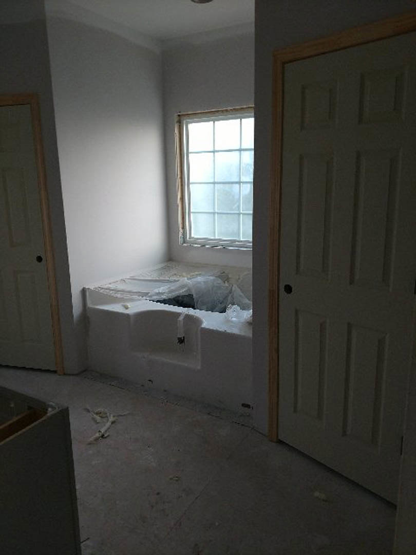Freestanding white bathtub on gray tile floor beside large window, plaster walls, wood door, and chrome fixtures in modern bathroom
