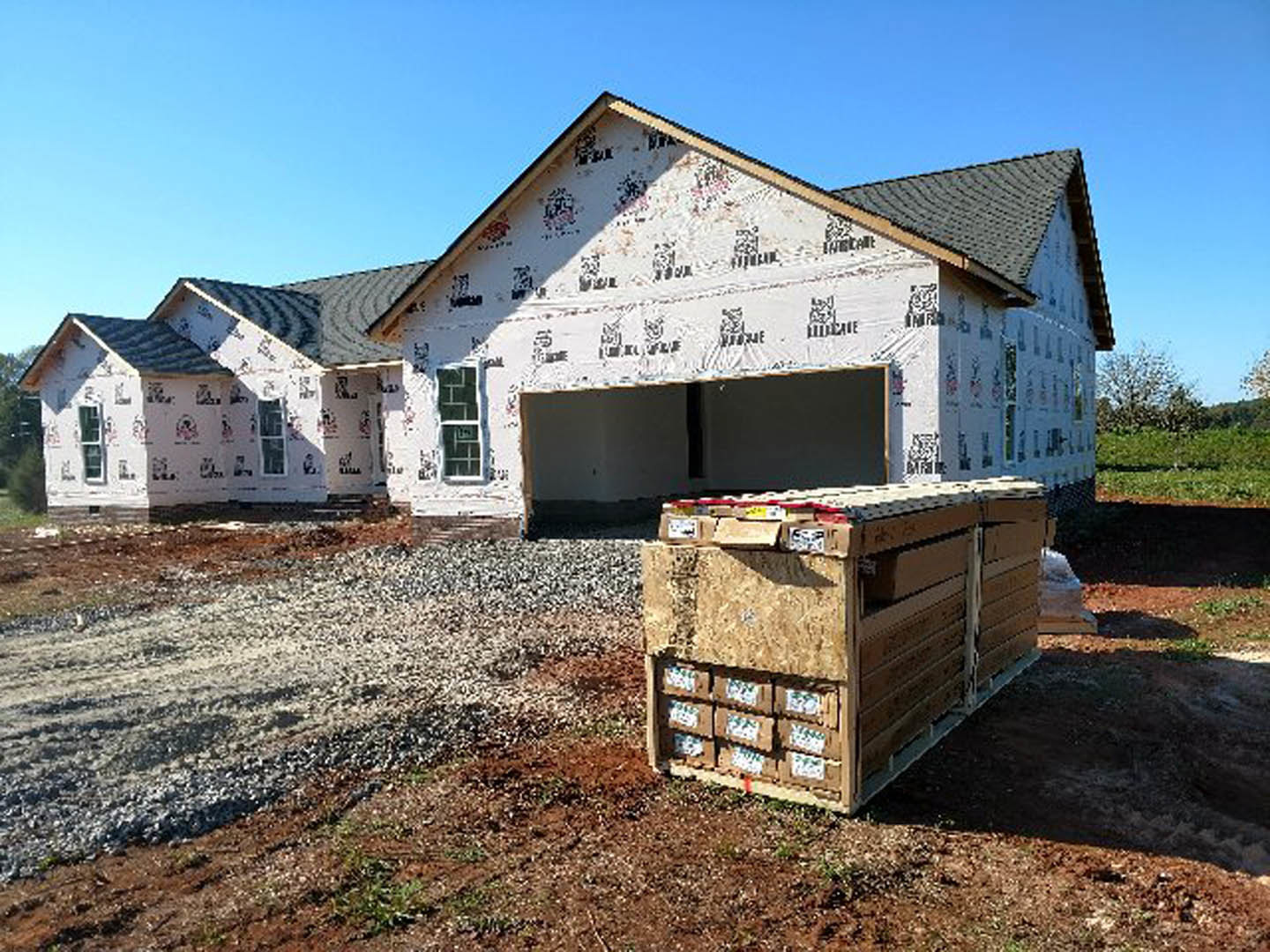 Partially built house with exposed framing, attached garage, stacked pallets of lumber and boxes on dirt lot, windows installed, blue sky overhead