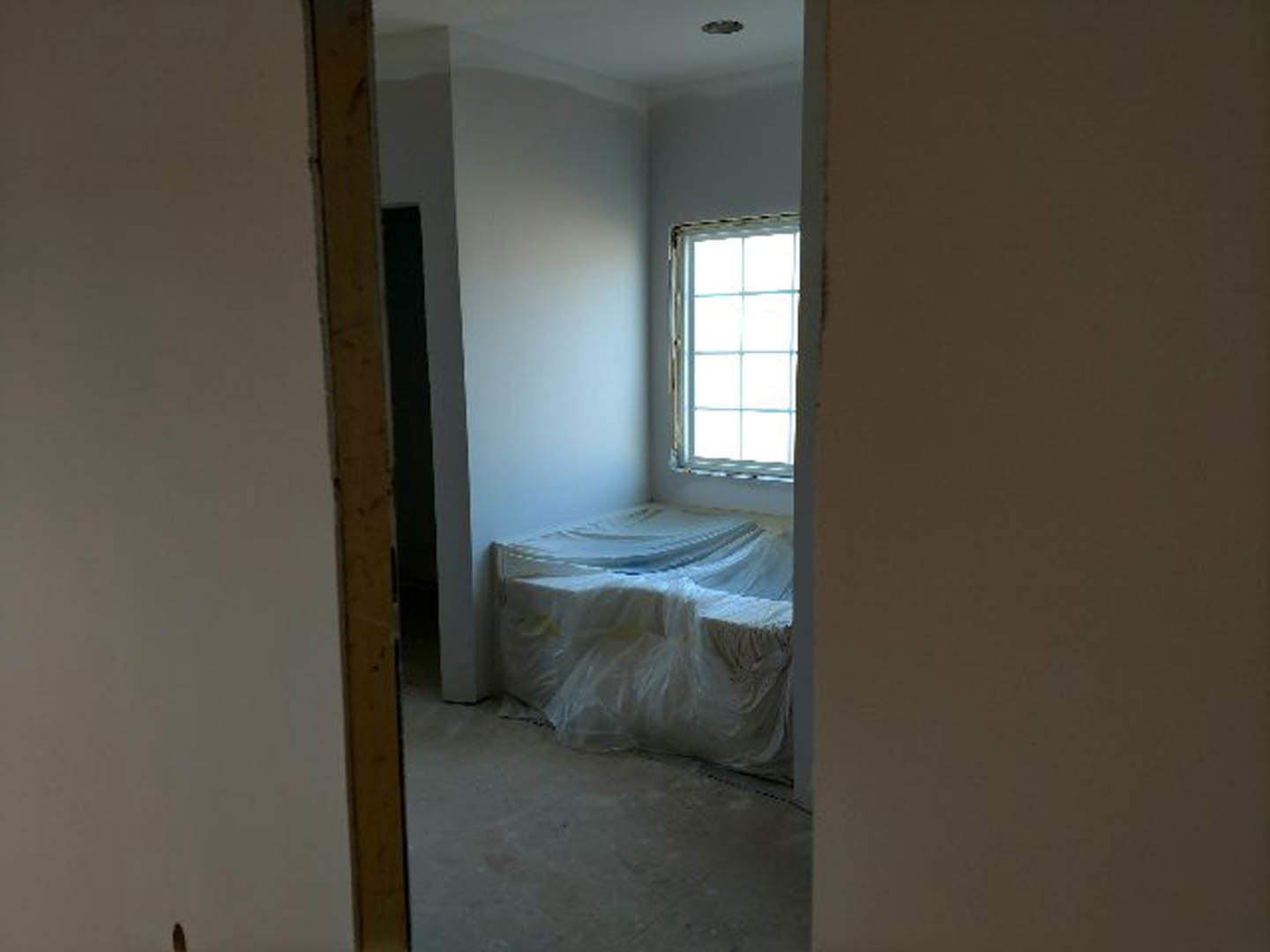 Bedroom under construction with bed wrapped in clear plastic, white-framed window, white walls, exposed wooden post, and bag of sand on unfinished floor