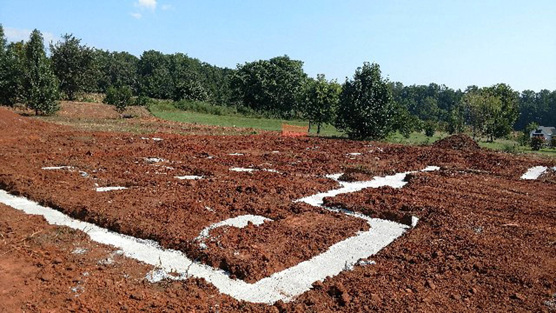 Dirt field marked with white lines, surrounded by grass and trees under a cloudy sky
