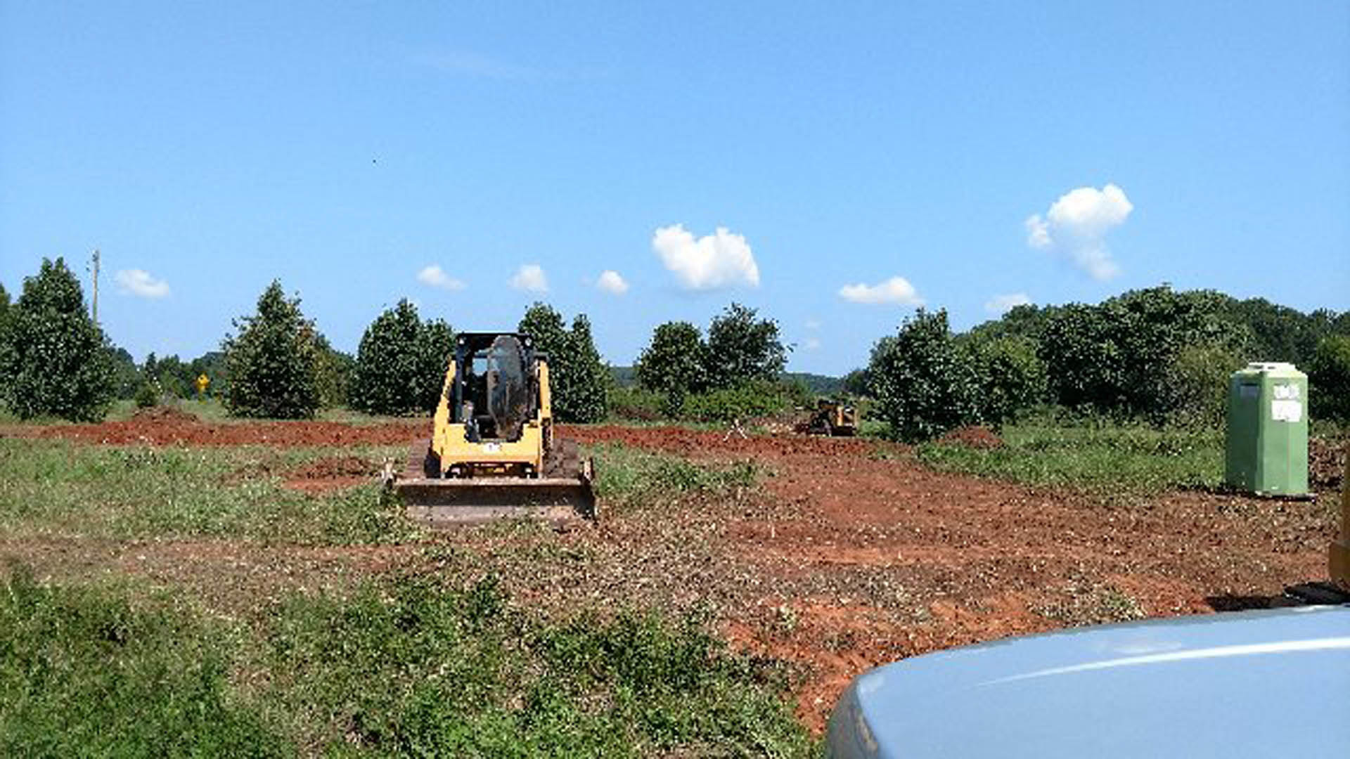 Yellow bulldozer parked on grassy, soil-covered lot with scattered trees and cloudy sky in background