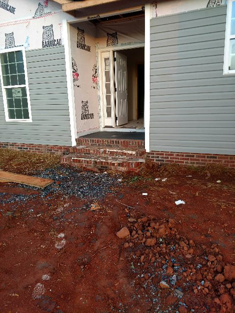 White house exterior with open door, red dirt and rocks piled near entry, window displaying sign, wood plank leaning against siding