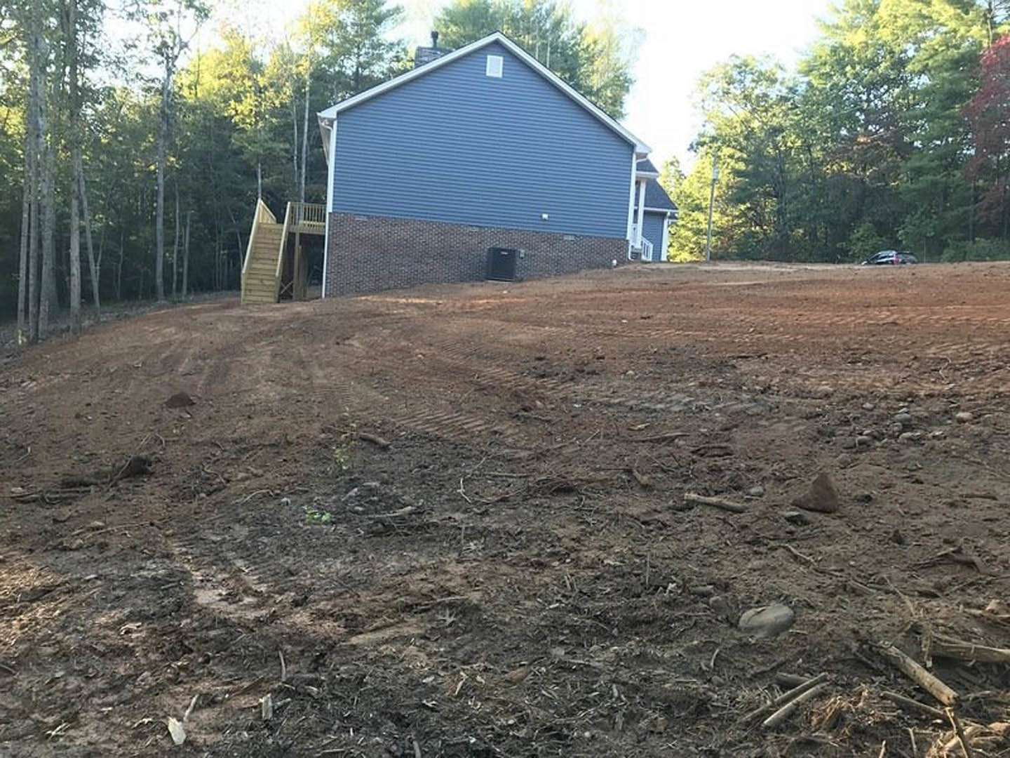 Two-story house with brick chimney and brick wall, surrounded by a large dirt hill and sparse grass, trees in the background under a clear sky