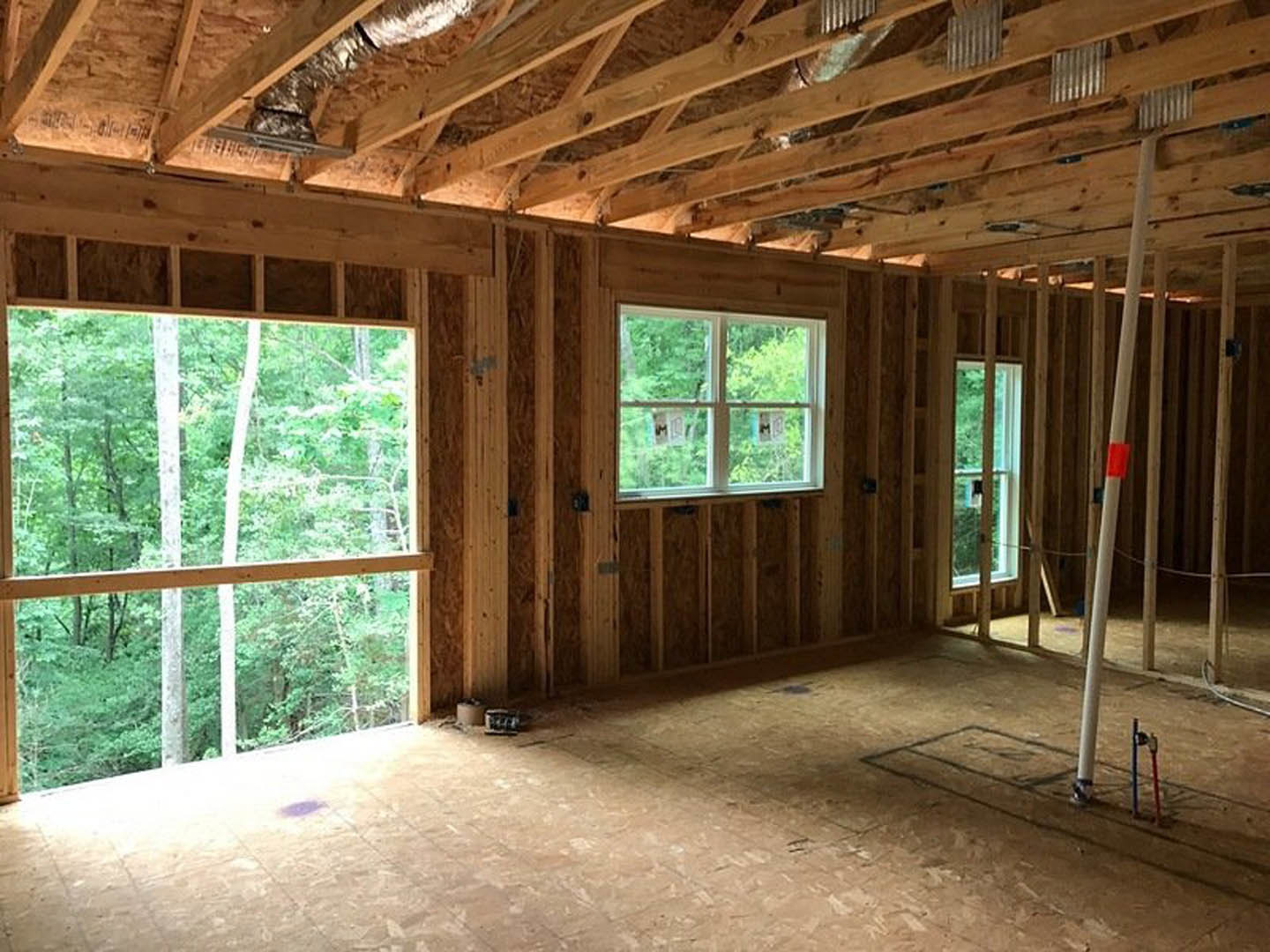 Sunlit room with large windows, exposed wooden beams, wood plank flooring, and a ladder leaning against the wall; trees visible through the windows.