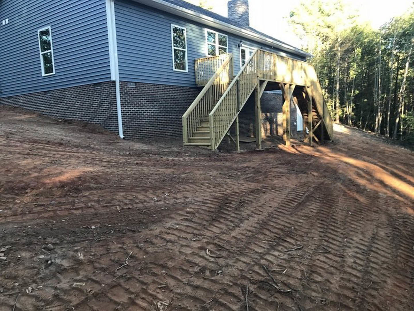 Two-story house with white-framed windows, wooden deck and staircase leading down a dirt hill, tire tracks visible on the ground, chimney partially visible against a blue sky.