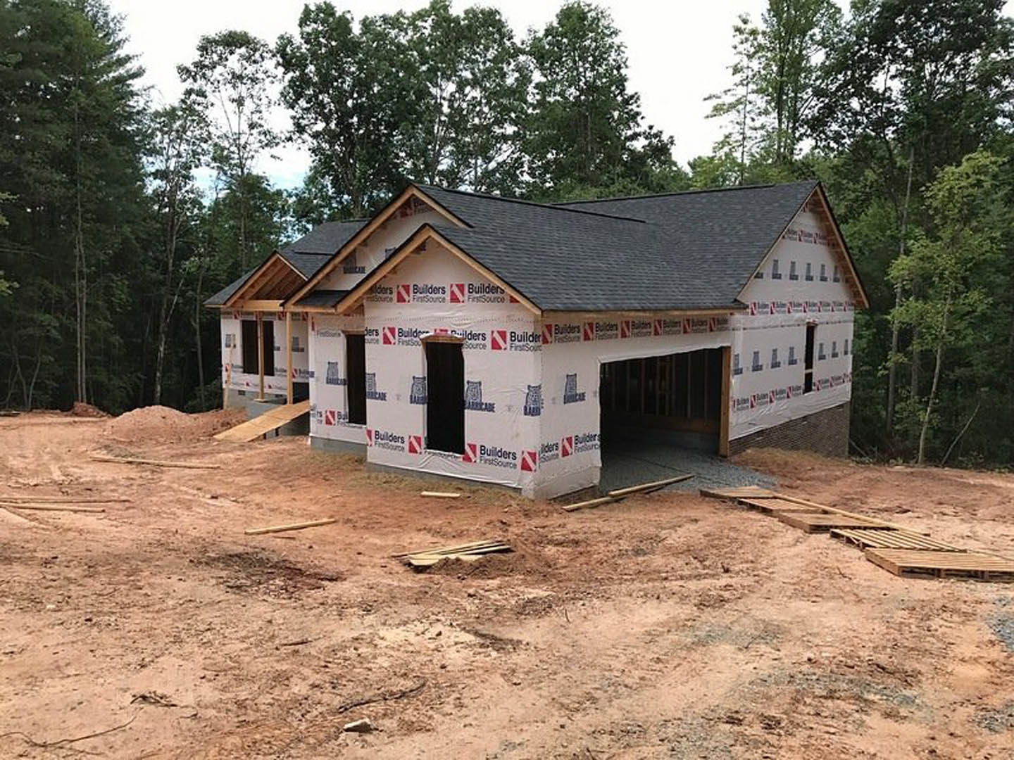 Wood-framed house under construction with exposed plywood sheathing, attached garage, surrounded by tall trees and dirt ground
