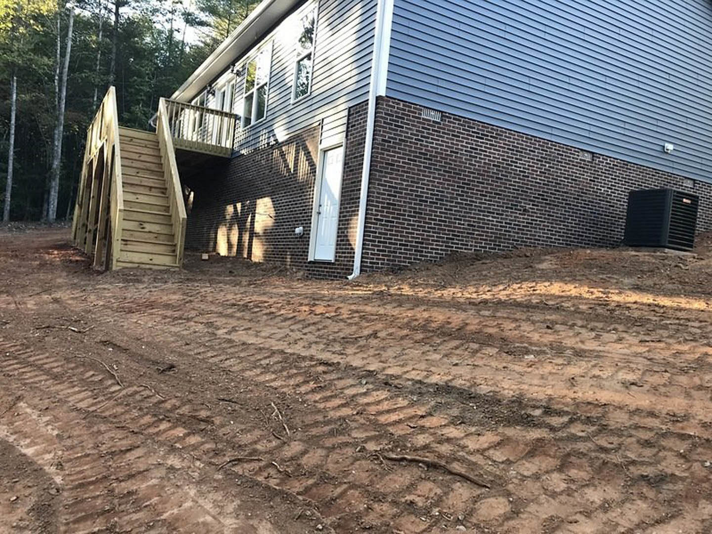 Brick house with white door, wooden exterior staircase, dirt road with tire tracks, fenced yard, trees and plants under blue sky