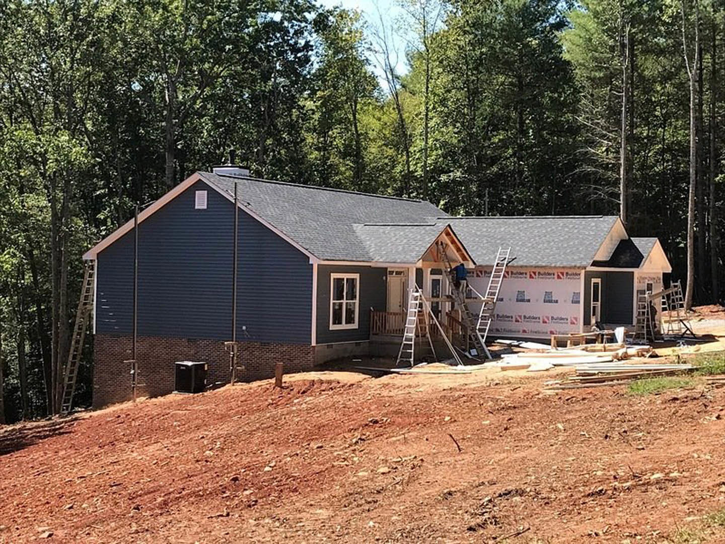 Partially built house with exposed framing, white window trim, ladders leaning against exterior, dirt mound in foreground, worker climbing ladder, surrounding trees in background