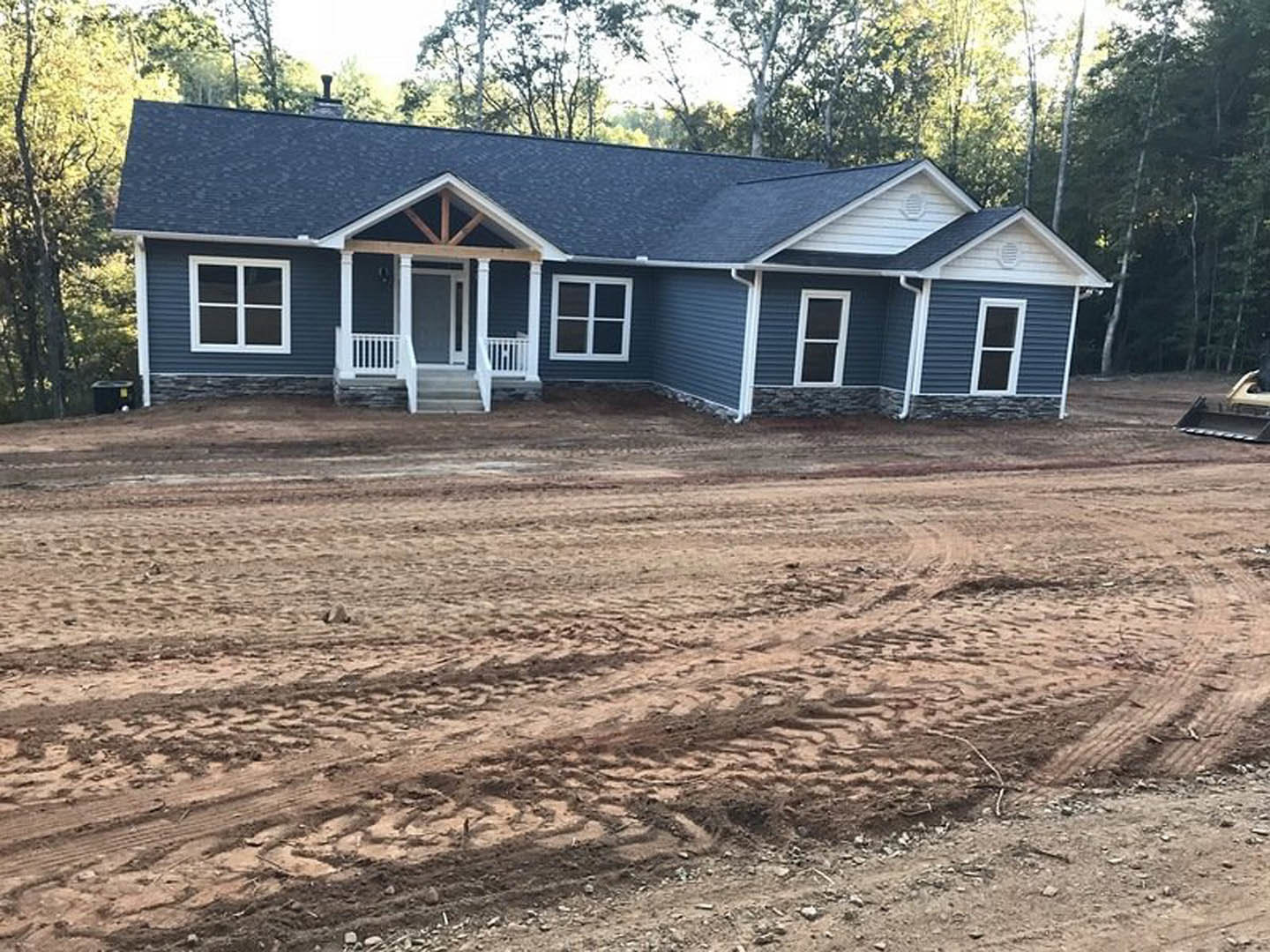 Two-story cottage-style home with light siding, white-framed windows, covered porch with white railing, dirt road with tire tracks leading to entrance, surrounded by mature trees