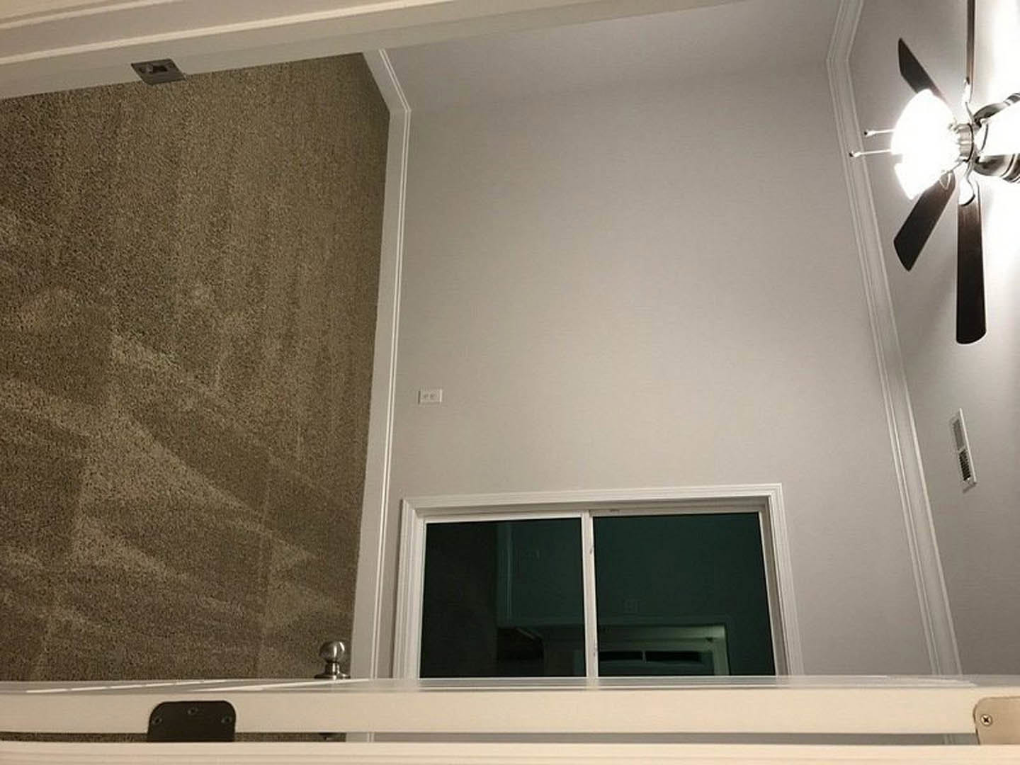 White paneled door with brushed metal knob, beige carpet flooring, white plaster walls, ceiling fan with dark blades, window with black-out curtain