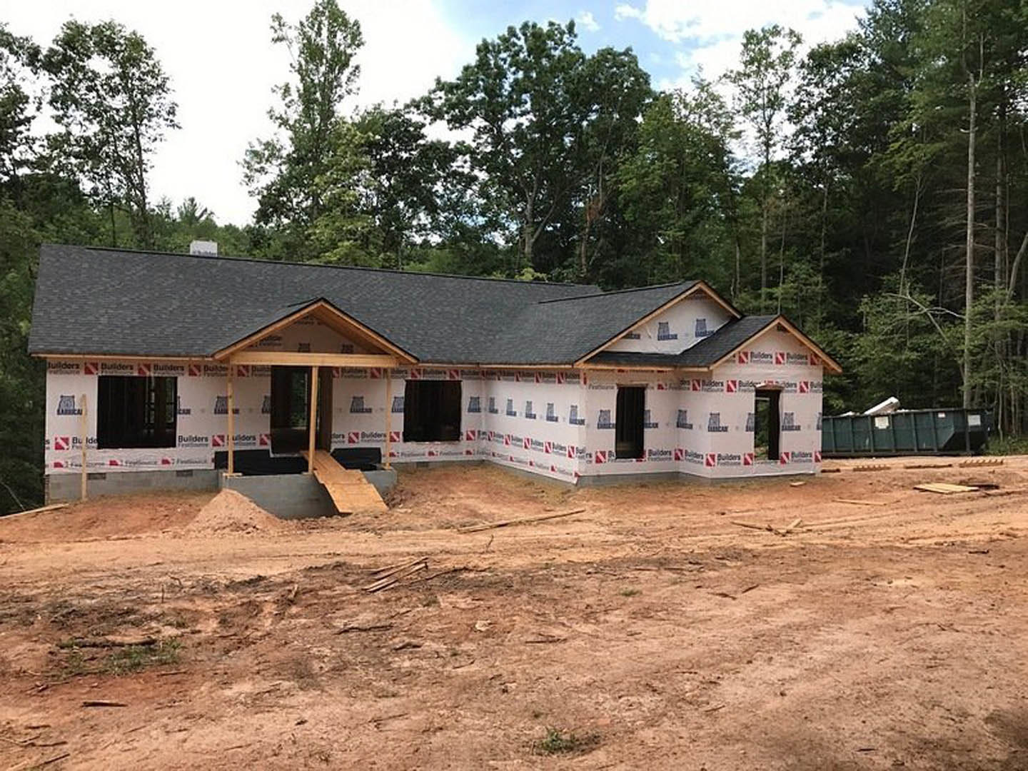 Wood-framed house under construction surrounded by tall trees, exposed beams and unfinished exterior, dirt lot in foreground, blue sky with scattered clouds