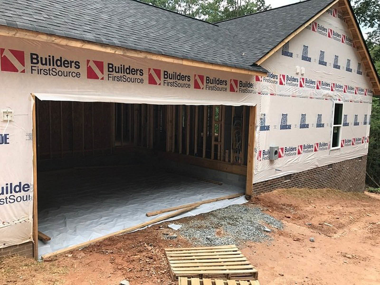 Partially built house with exposed framing, brick garage wall, square-paned window, wood pallets on rocky ground, dirt and tarp, red and white construction sign.