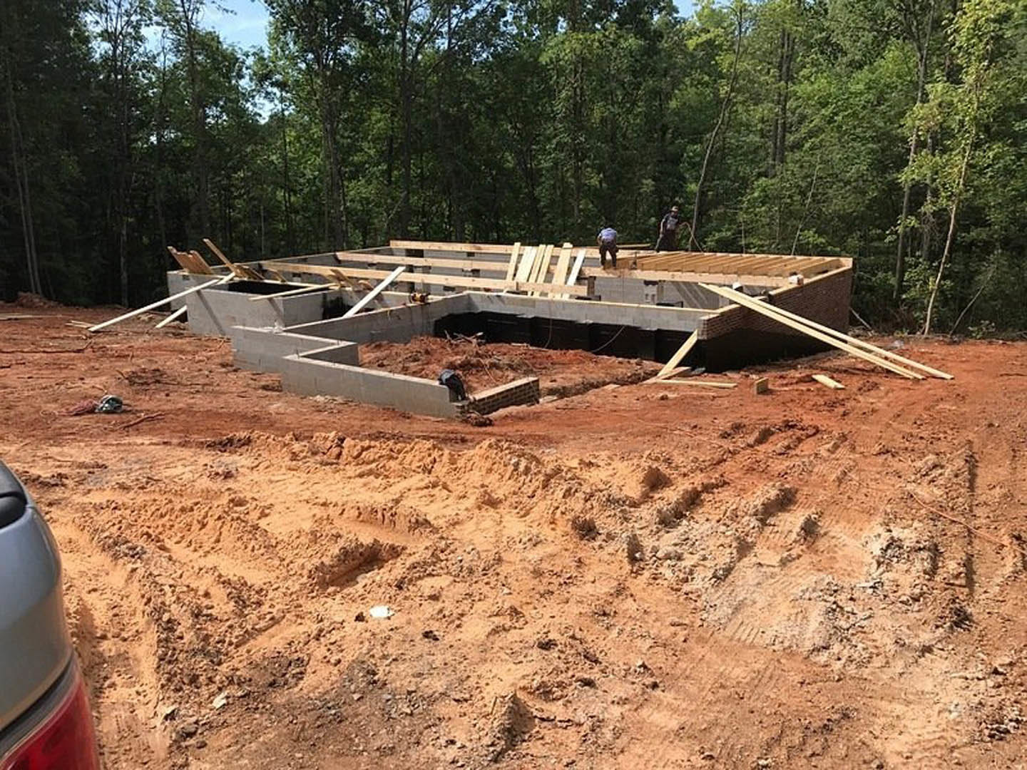 Wood-framed house under construction with workers assembling beams, exposed foundation, dirt ground, scattered lumber, and trees in the background