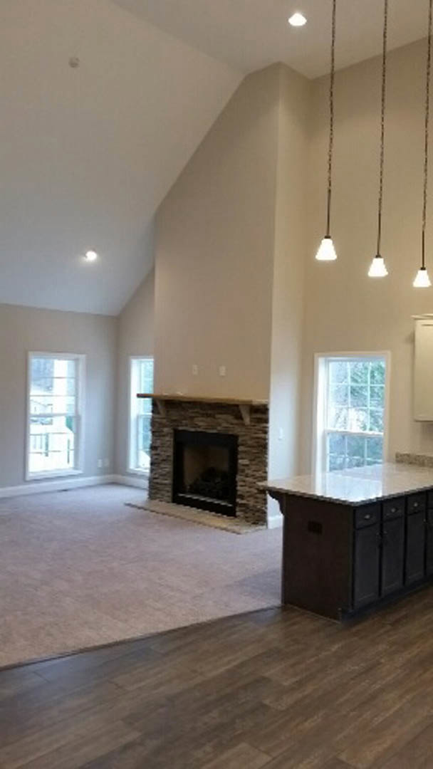 Open-concept living room with hardwood floors, stone fireplace featuring a wood mantel, white walls, large window with white trim, and kitchen island with built-in drawers