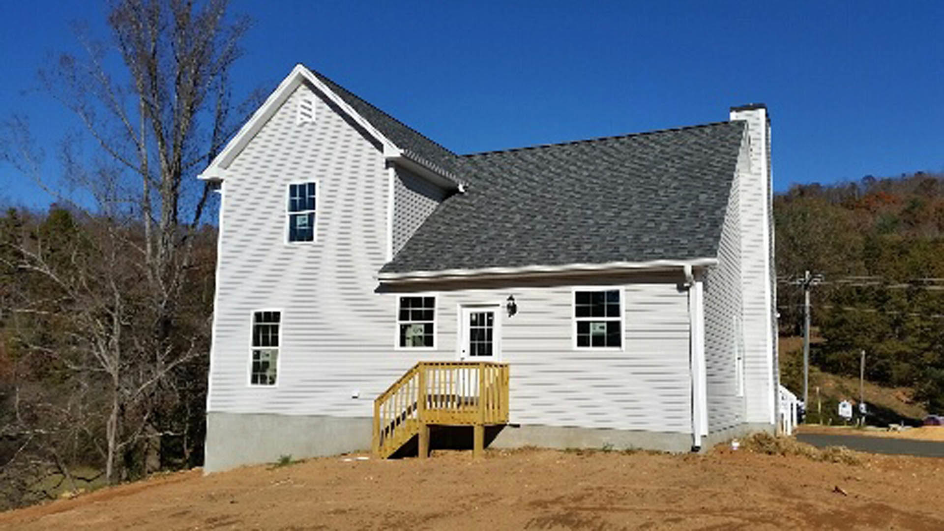 Two-story house with light siding, covered front porch, wooden stairs with railings, leafless tree nearby, dirt ground, and shingled roof