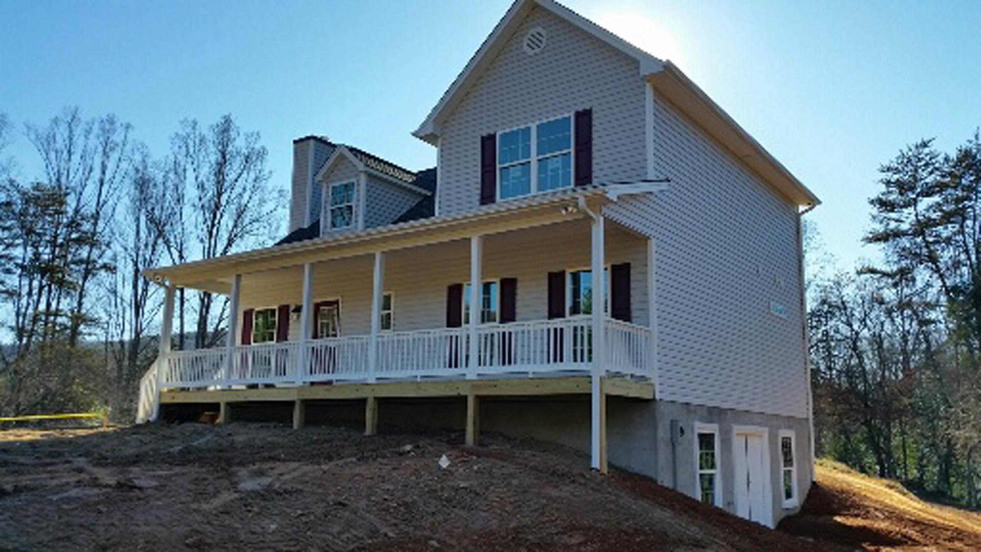Two-story house with white siding and porch, white railing, situated on a grassy hill with dirt patches, surrounded by trees under a blue sky