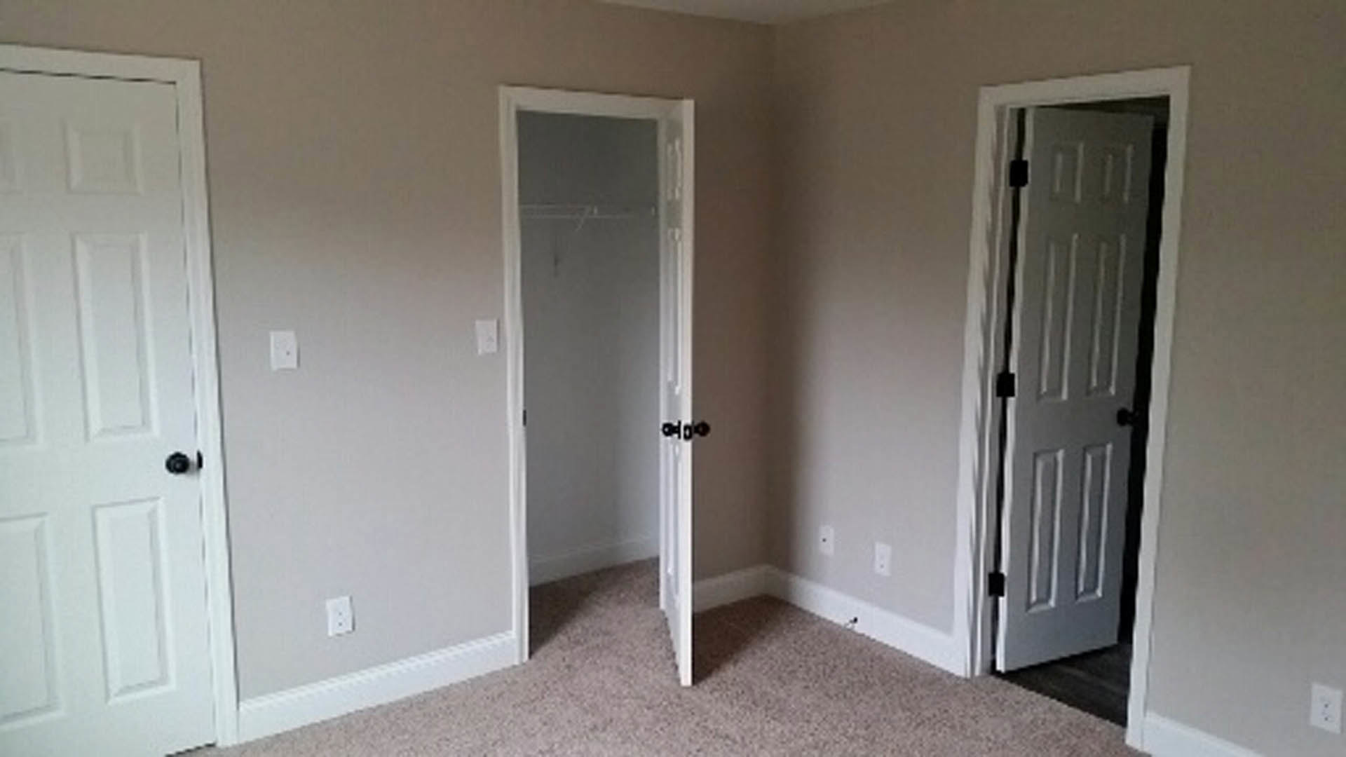 White closet doors with brushed metal handles set against light gray walls, hardwood flooring, and minimal decor in a modern bedroom.