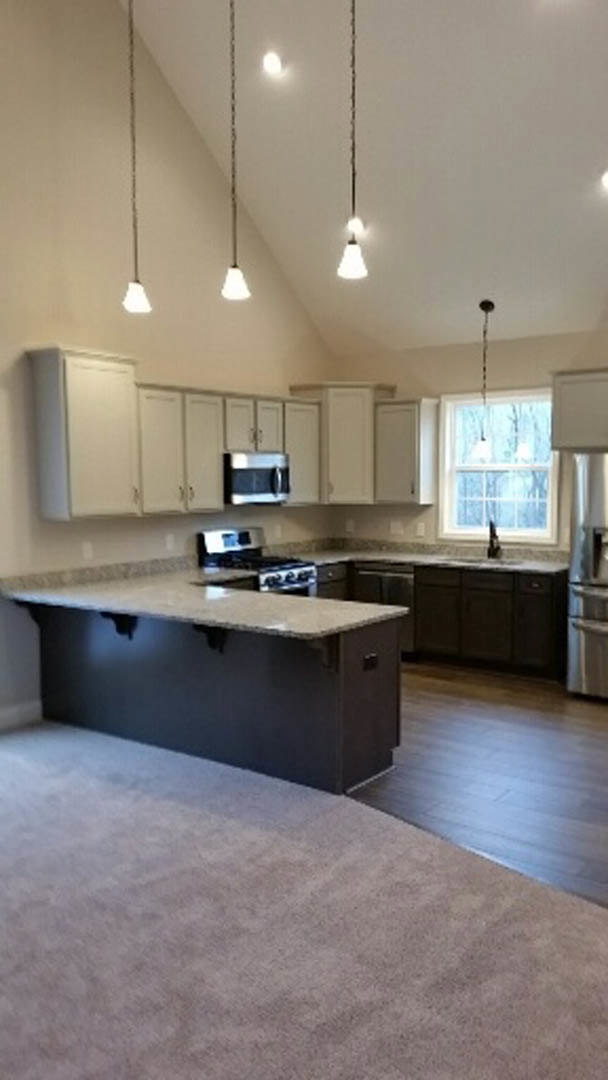 Spacious kitchen featuring a large central island with stone countertop, stainless steel stove, white cabinetry, row of pendant lights, and window with white trim.