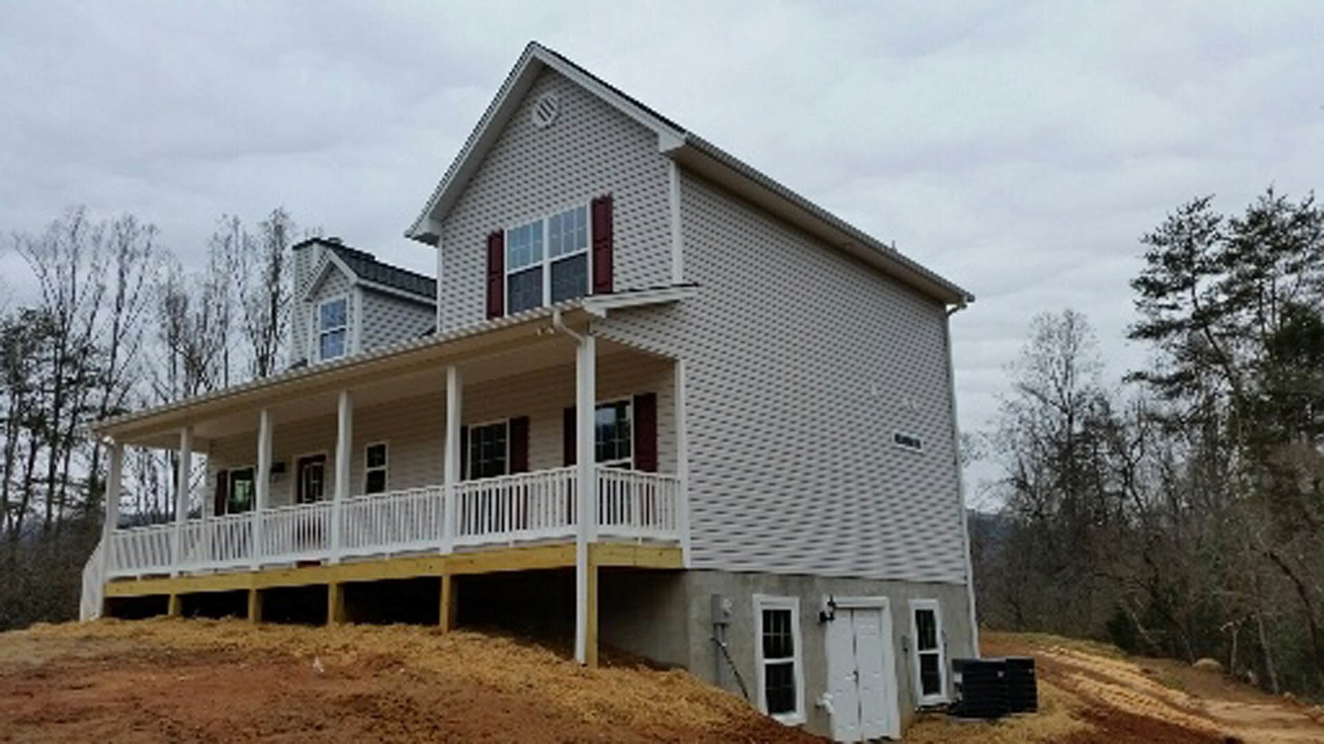 Two-story house with gray siding, white porch railing, large windows, and a covered front porch, situated on a grassy hill with trees in the background