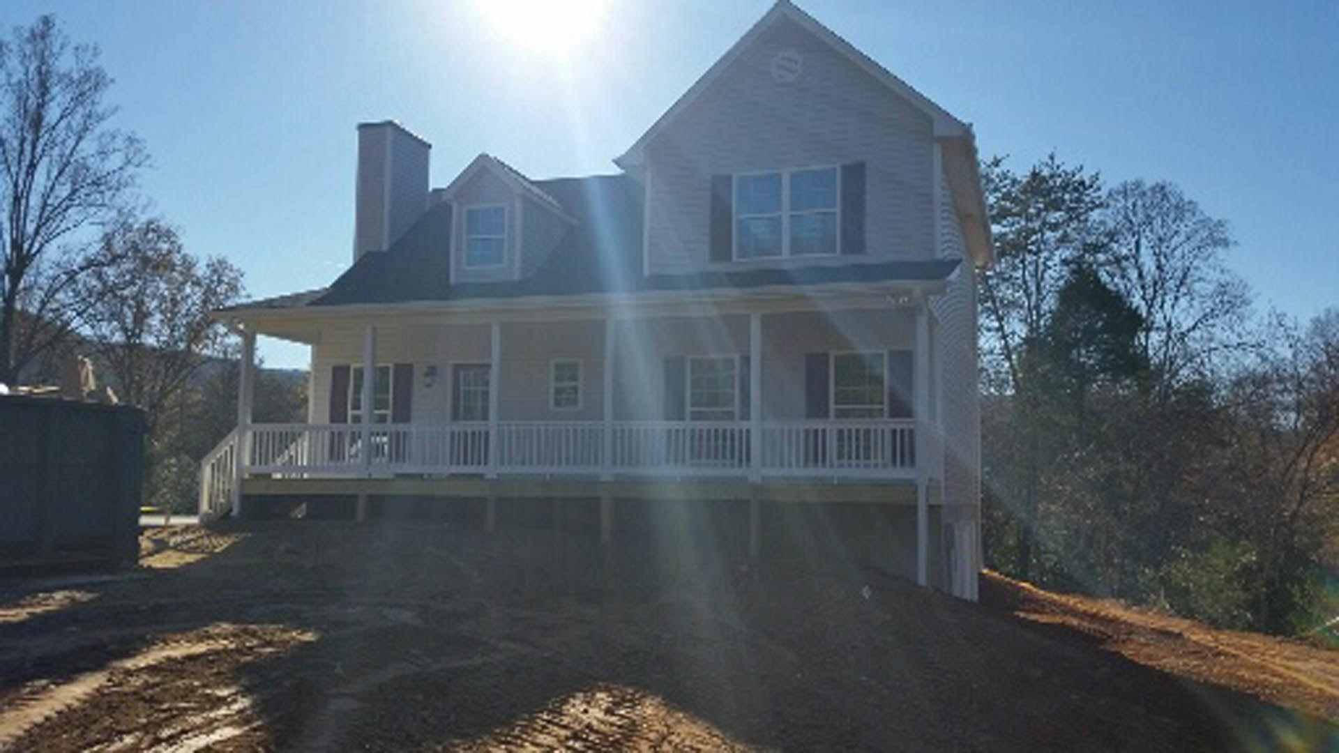 Two-story home with white porch railing, grid windows, and large covered porch set on a grassy hill with trees in the background