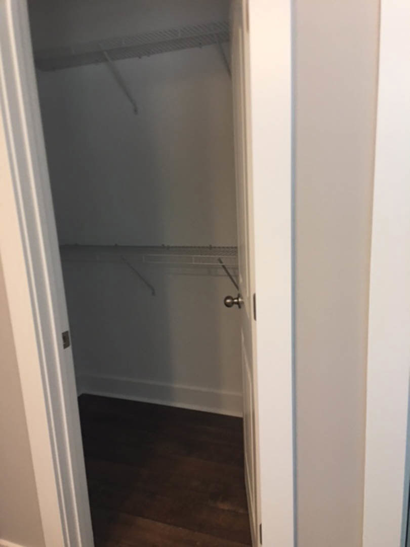 Open white closet door with black handle, revealing metal shelf and dark wood flooring, blurred hand partially visible.