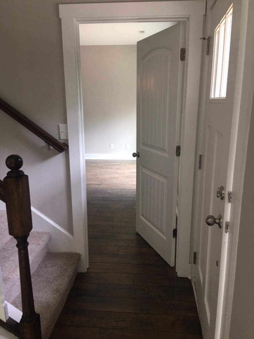 Hallway with light wood flooring, white plaster walls, a wooden staircase with vertical posts, and a closed door featuring a metal handle