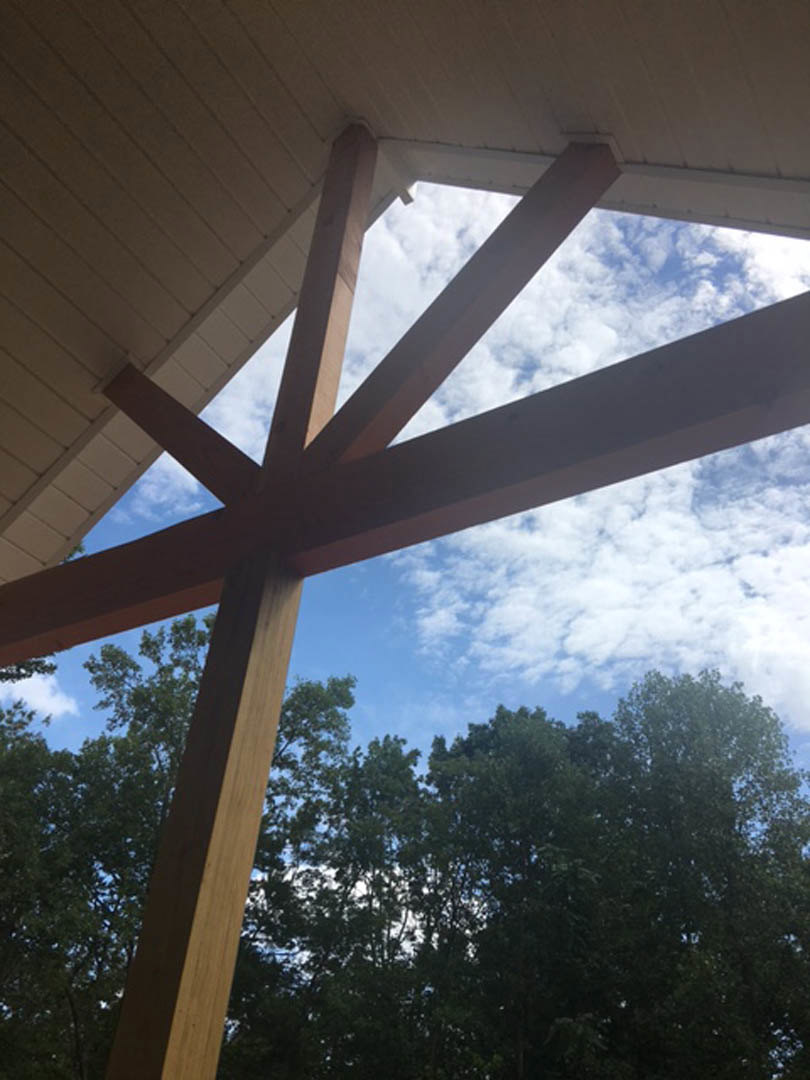 Exposed wood beams supporting a roof with natural wood finish, surrounded by leafy trees under a blue sky with scattered clouds