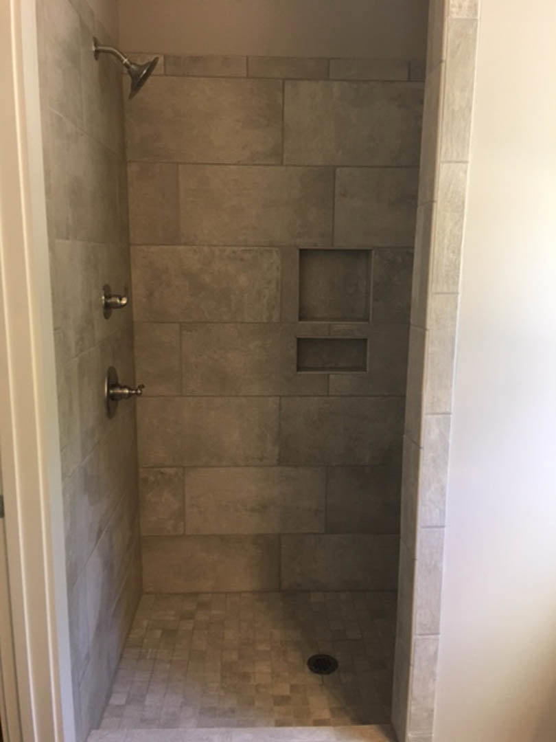 Walk-in shower featuring a tile floor and tile walls, chrome shower head mounted on a white wall with a white border, bathroom door visible in the background