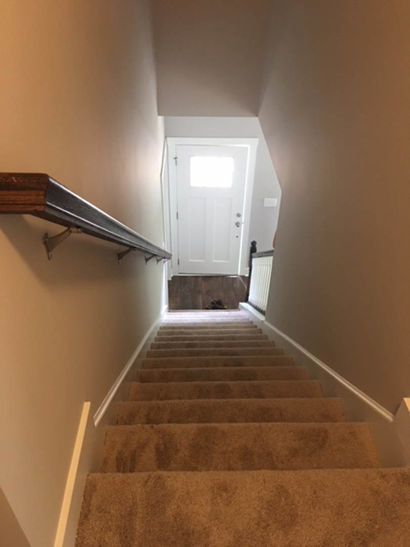 Carpeted staircase with wooden handrail ascending to a white door with glass window, wooden shelf mounted on plaster wall, blurred figure near wood accent wall