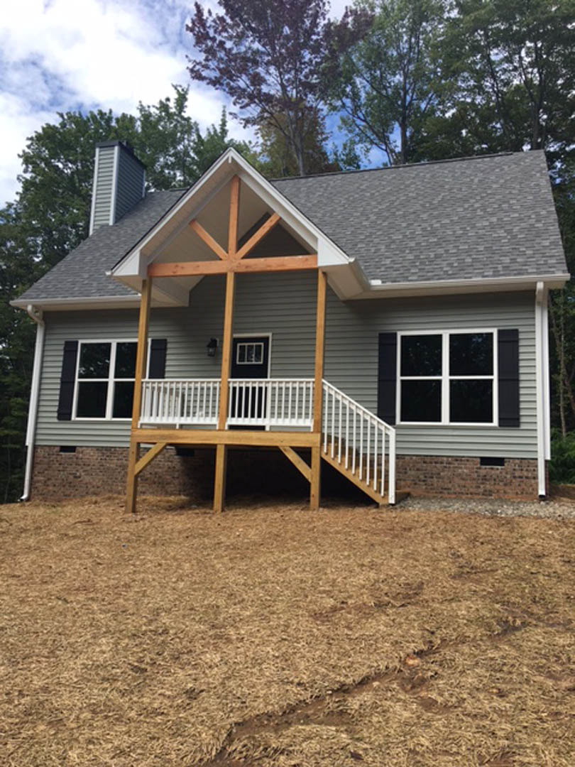 Two-story cottage-style home with gray siding, white-framed windows, and two covered porches featuring white railings, set beside a brown grass lawn and surrounded by trees under a