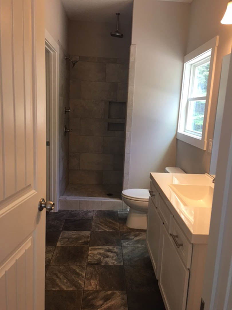 Modern bathroom featuring a white porcelain sink and toilet, gray tile flooring, glass-enclosed shower with textured tile walls, white-framed window, and brushed metal door knob