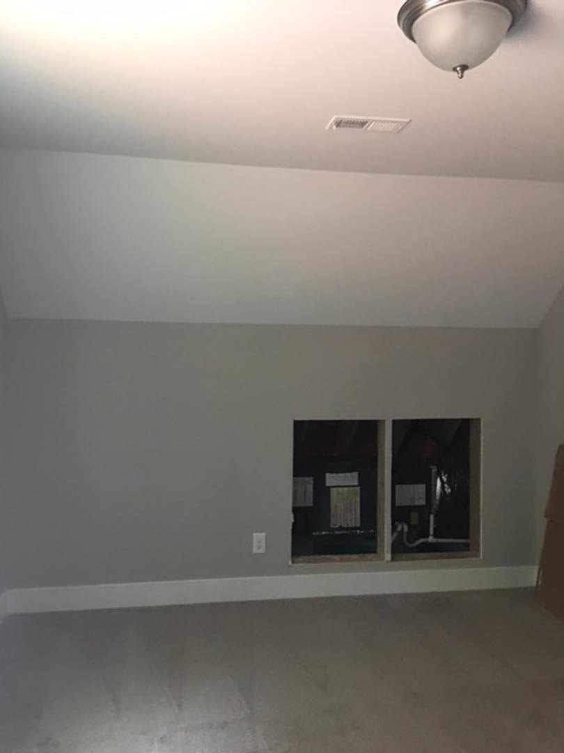 White-walled room with grey flooring, large window, black corner trim, ceiling fan, and person standing near window with visible pipe and paper